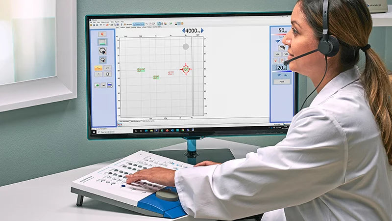 Audiologist operating a clinical audiometer beside a hearing test display